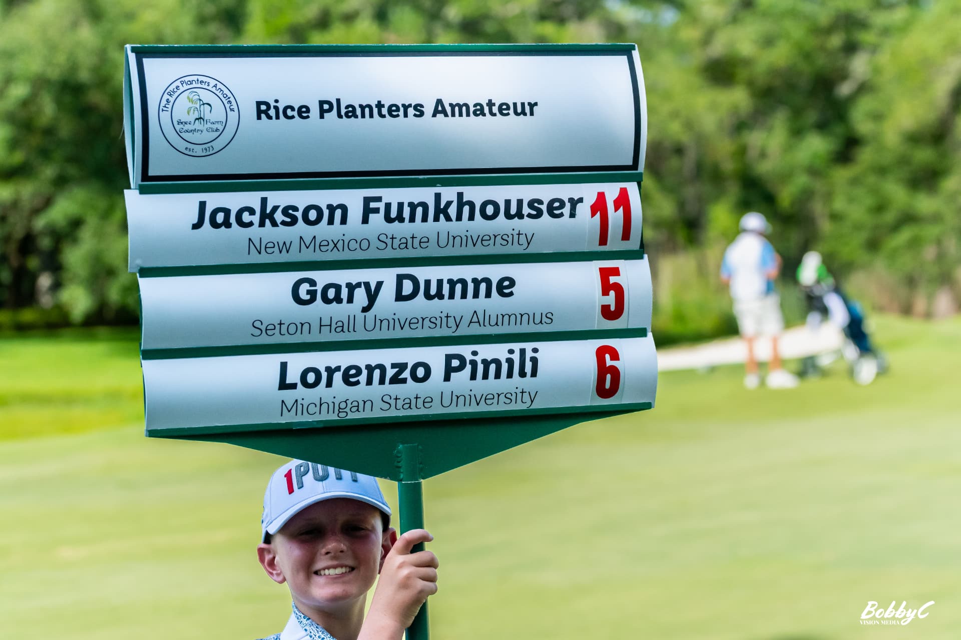 A young standard bearer smiling as he carries the Rice Planters Amateur scoreboard sign on course at Snee Farm